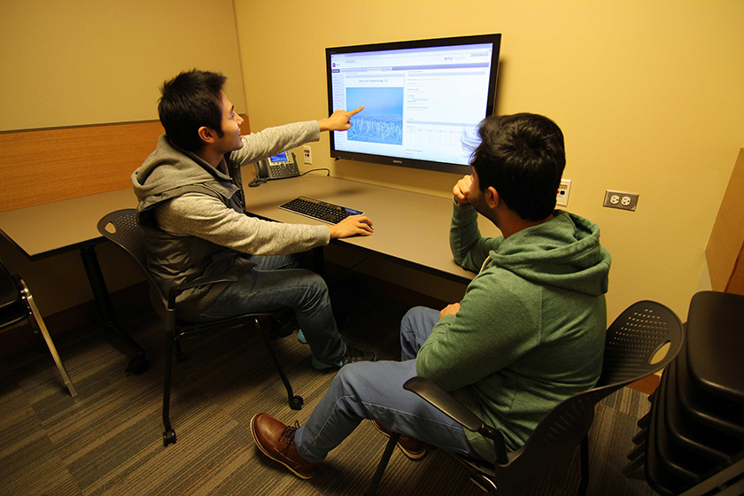 Two people sitting in a consultation room, one person is pointing to the computer screen mounted on the wall screen and controlling the mouse while the other looks on.